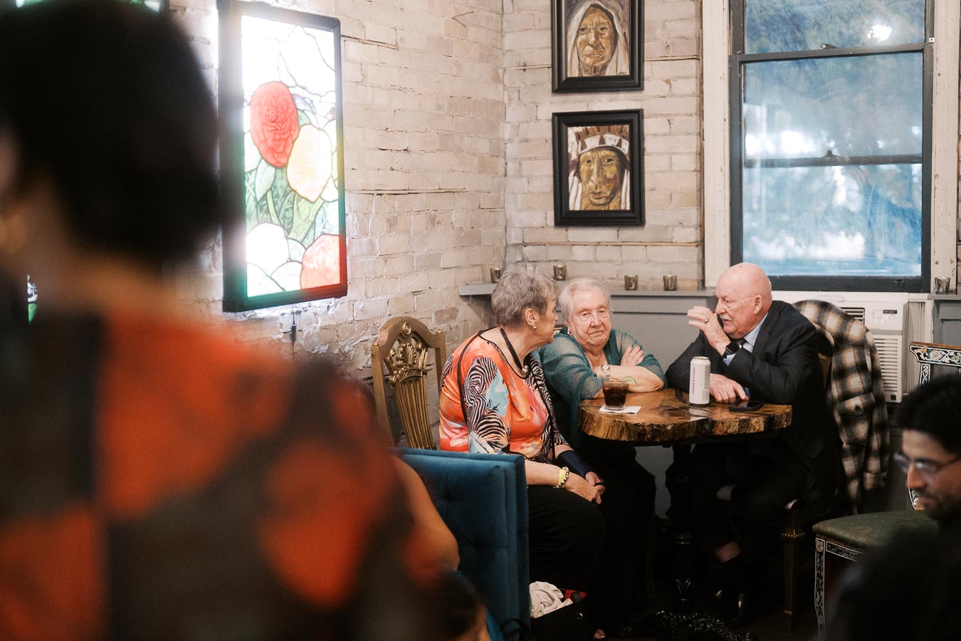 Elderly group sitting at a rustic cafe table, engaging in conversation. The cozy setting features brick walls, vibrant stained glass art, and portraits, creating a warm atmosphere.