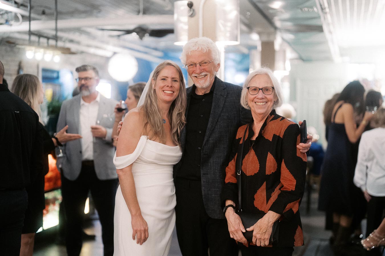 A woman in a white dress stands with two older adults, smiling, at a lively indoor event, with a blurred background of people socializing.