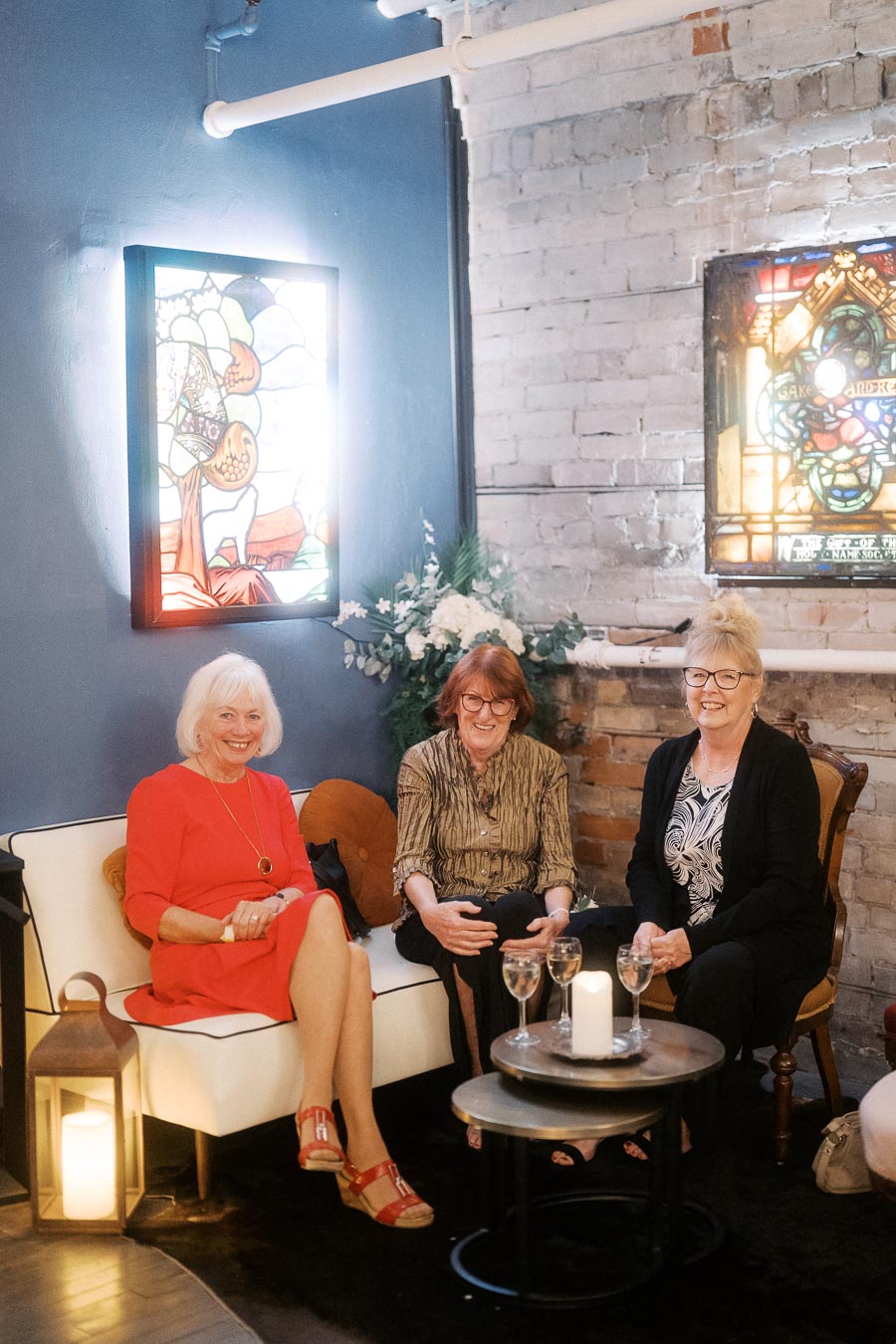 Three women enjoying a cozy, stylish gathering in a lounge with exposed brick walls and stained glass decor, seated around a small table with glasses of wine.