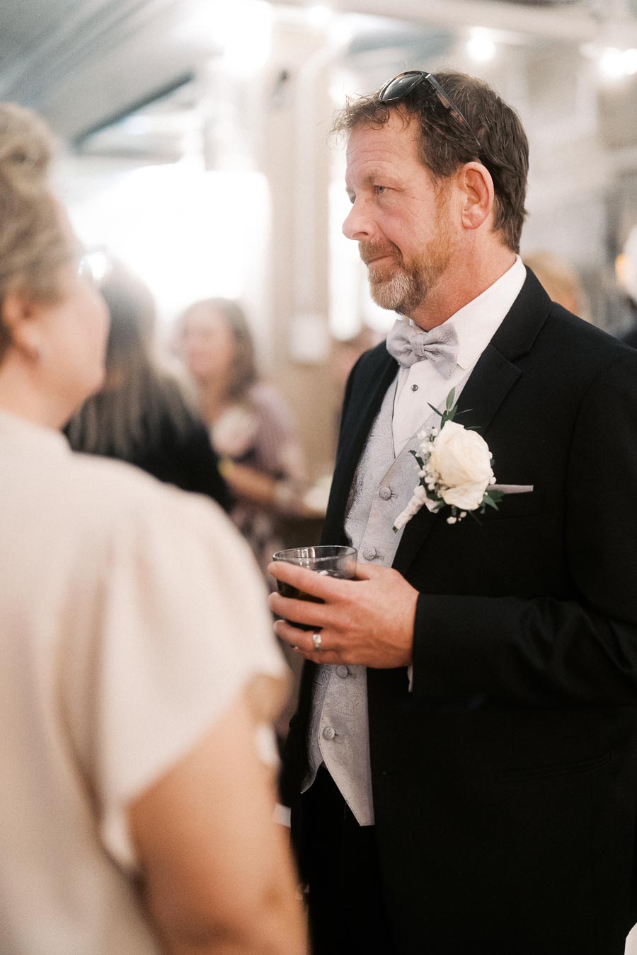 A man in a formal suit with a bow tie and boutonniere, holding a drink, converses with another guest at a social event. The background is softly blurred with people mingling in a well-lit venue.
