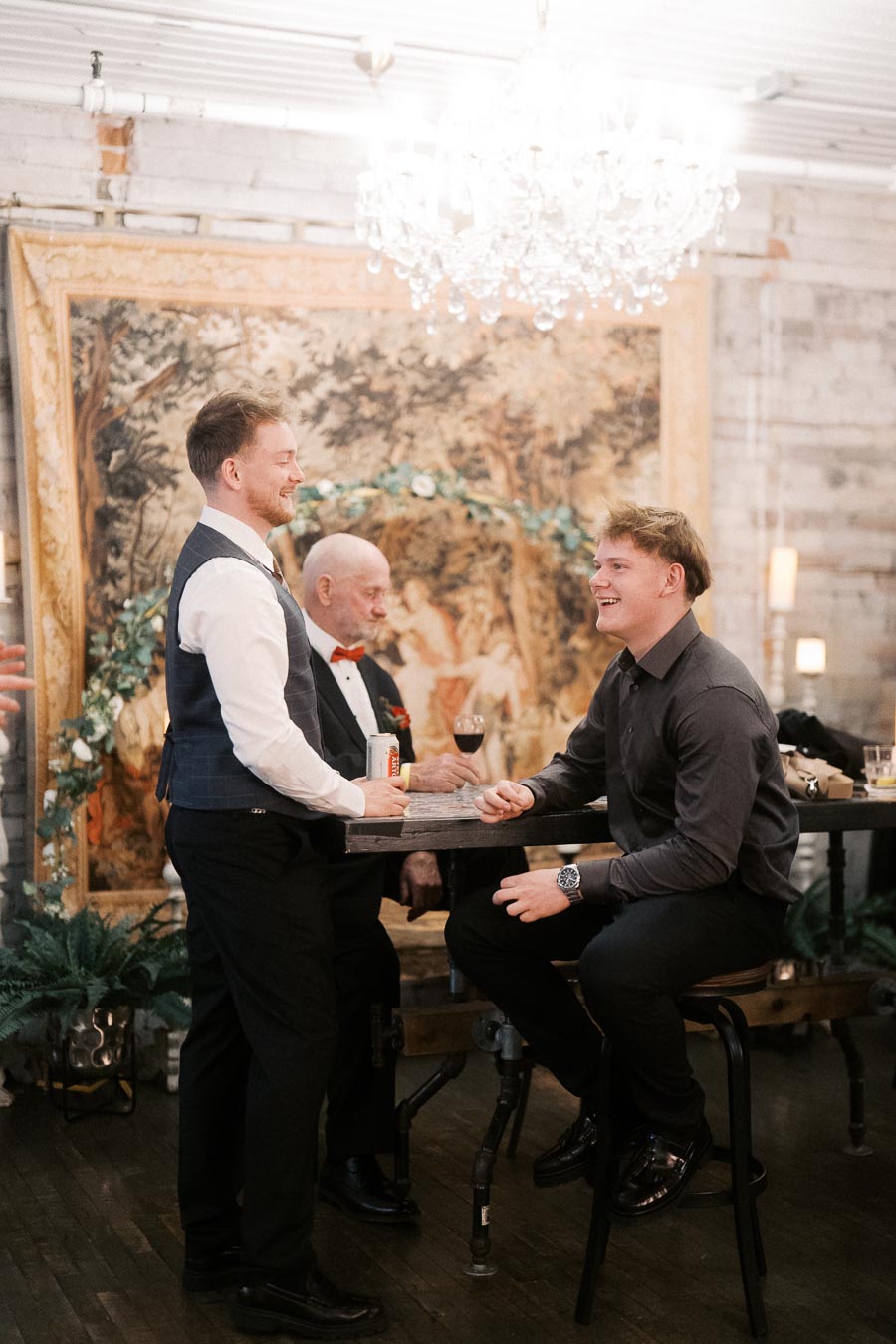 Group of people socializing at a wedding reception, elegantly dressed, with a vintage tapestry and chandelier in the background.