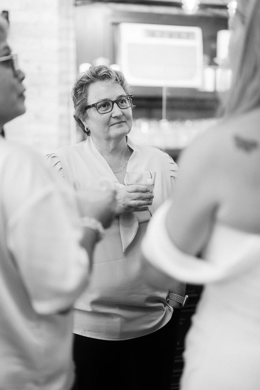 Black and white image of a group of people socializing, with a woman in glasses holding a drink in a casual gathering setting.