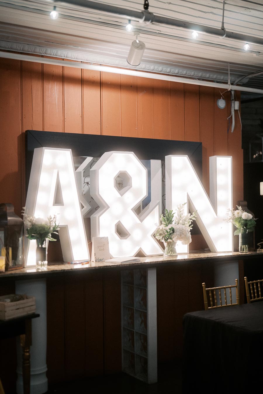 Illuminated marquee letters A & N displayed on a countertop with floral arrangements, set against a wooden wall backdrop in a dimly lit event space.