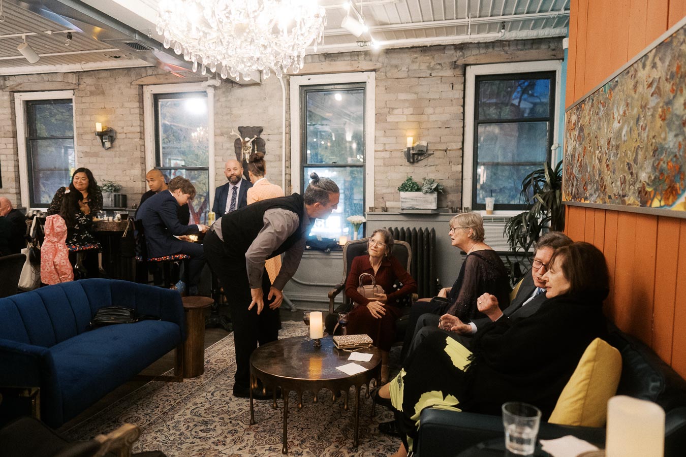 A cozy, warmly lit gathering in a stylish living room with people engaged in conversation, featuring a blue couch, patterned rug, and decorative wall art; a chandelier adds elegance to the ambiance.