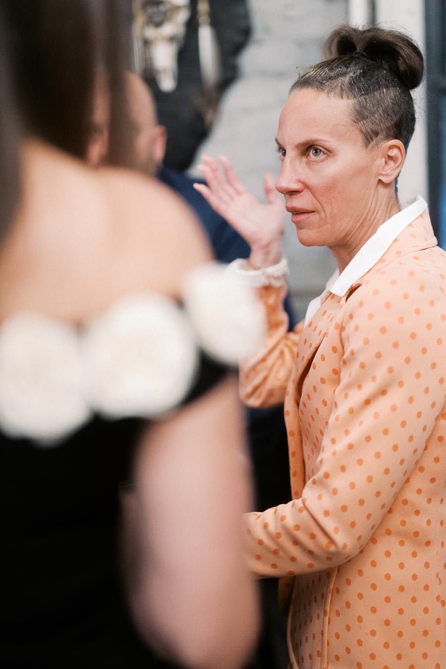 Woman in orange polka dot suit gesturing while conversing at social event.
