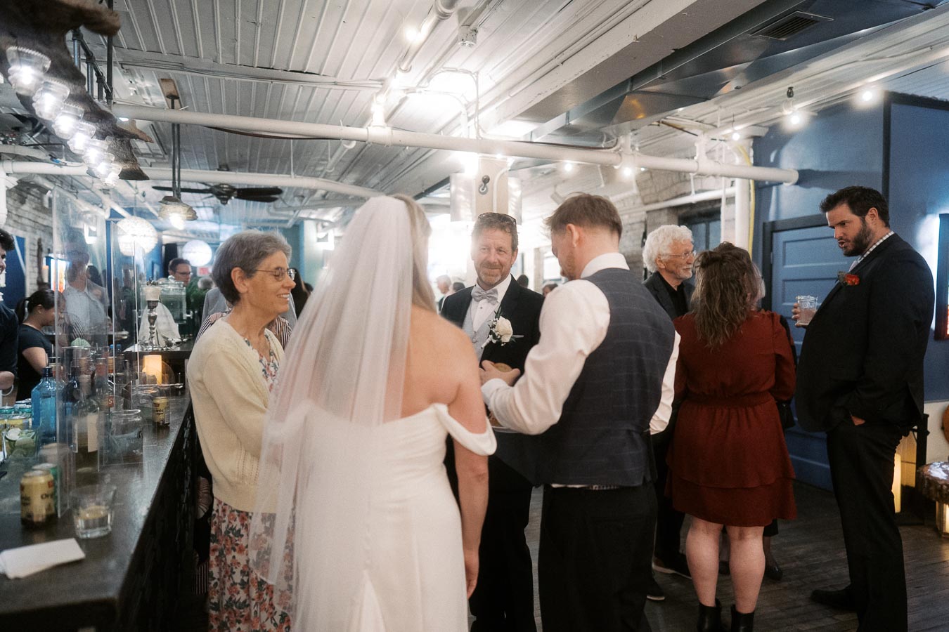 Guests mingle at a wedding reception in a cozy, dimly lit venue with exposed beams and hanging lights, featuring a bride in a white dress with a veil, a groom in a vest, and attendees in formal attire.