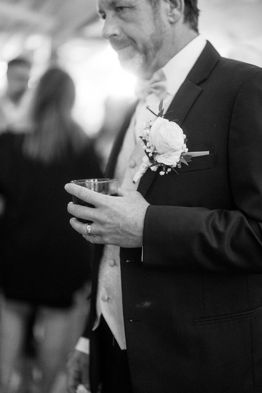 A black and white image of a man in formal attire at a wedding, holding a drink with a floral boutonniere pinned to his suit jacket.