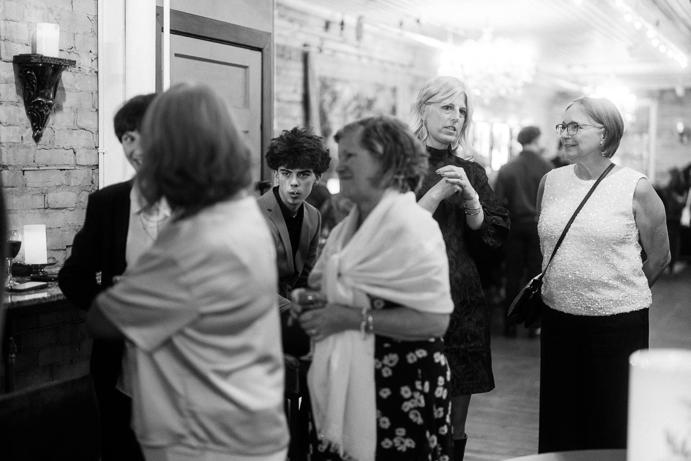 Black and white image of a social gathering with people conversing in a well-lit room; features a brick wall, candles, and elegantly dressed individuals creating a sophisticated atmosphere.