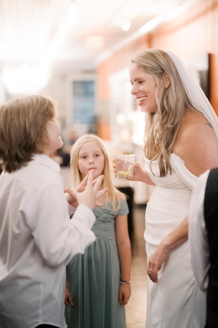 Bride smiling and chatting with two children at a wedding reception, holding a drink in a warmly lit room.