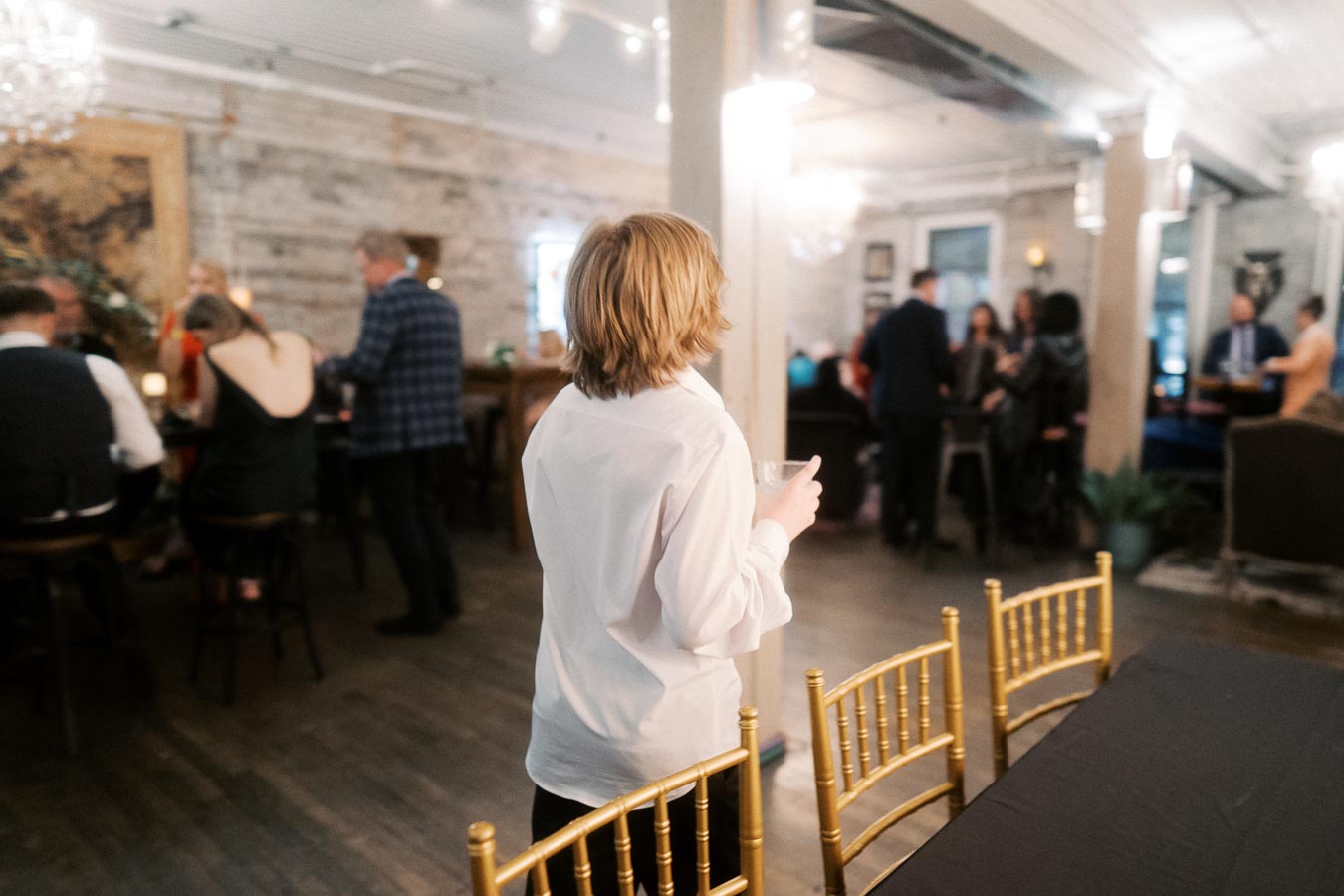 A young person in a white shirt holding a drink at a social gathering in a warmly lit event space with people mingling in the background.