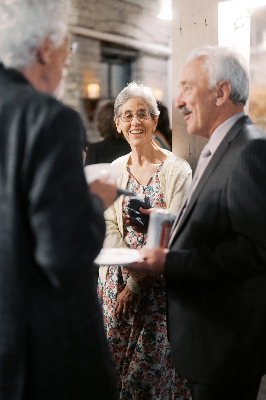 Elderly individuals enjoying a social gathering, engaged in a cheerful conversation, with one woman smiling and two men interacting, all in a warmly lit indoor setting.