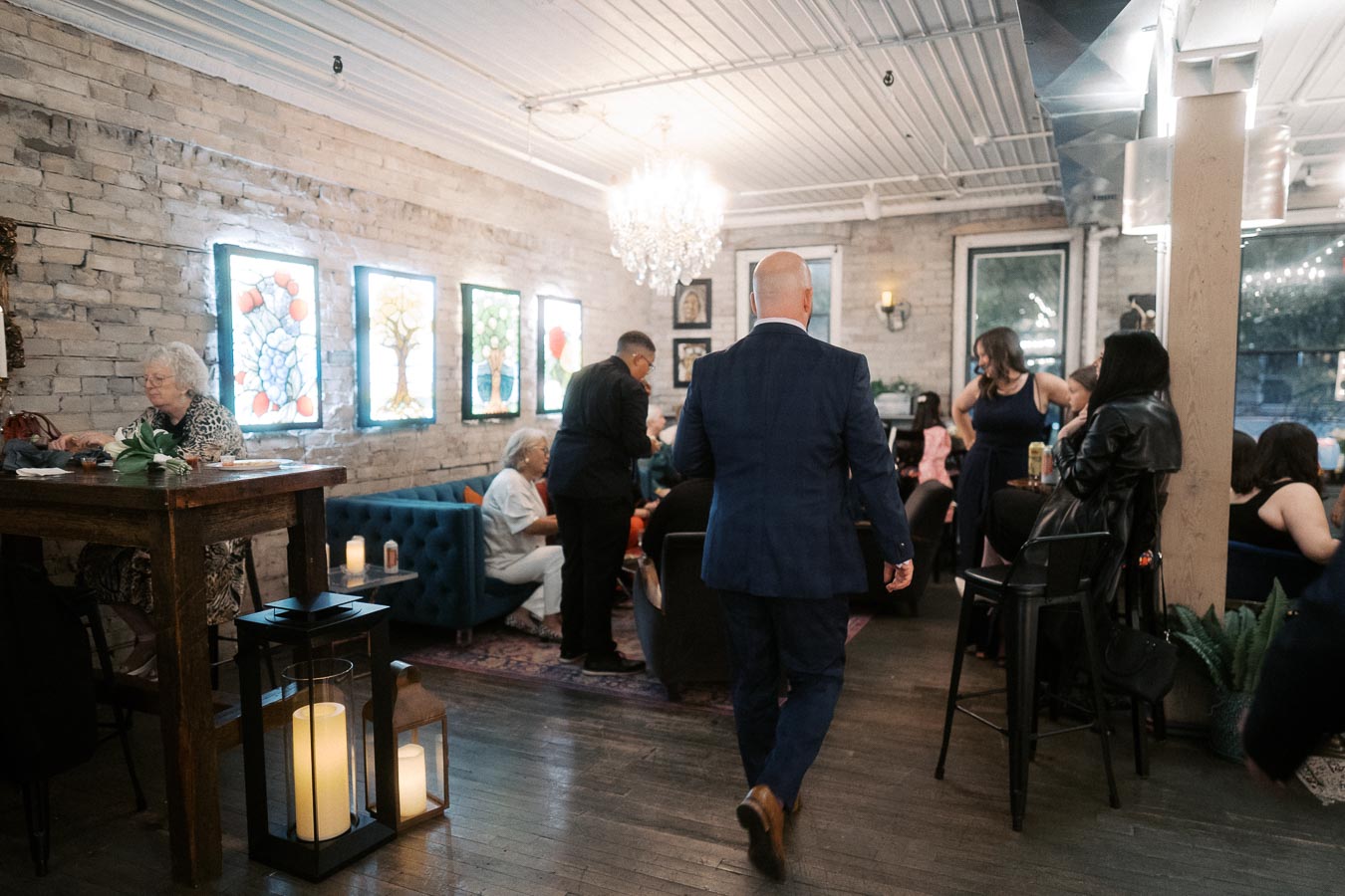 Elegant social gathering at a chic venue featuring exposed brick walls, stained glass art, and ambient lighting. Mixed age groups interact warmly around a table, with a focus on a man in a blue suit walking through the room.