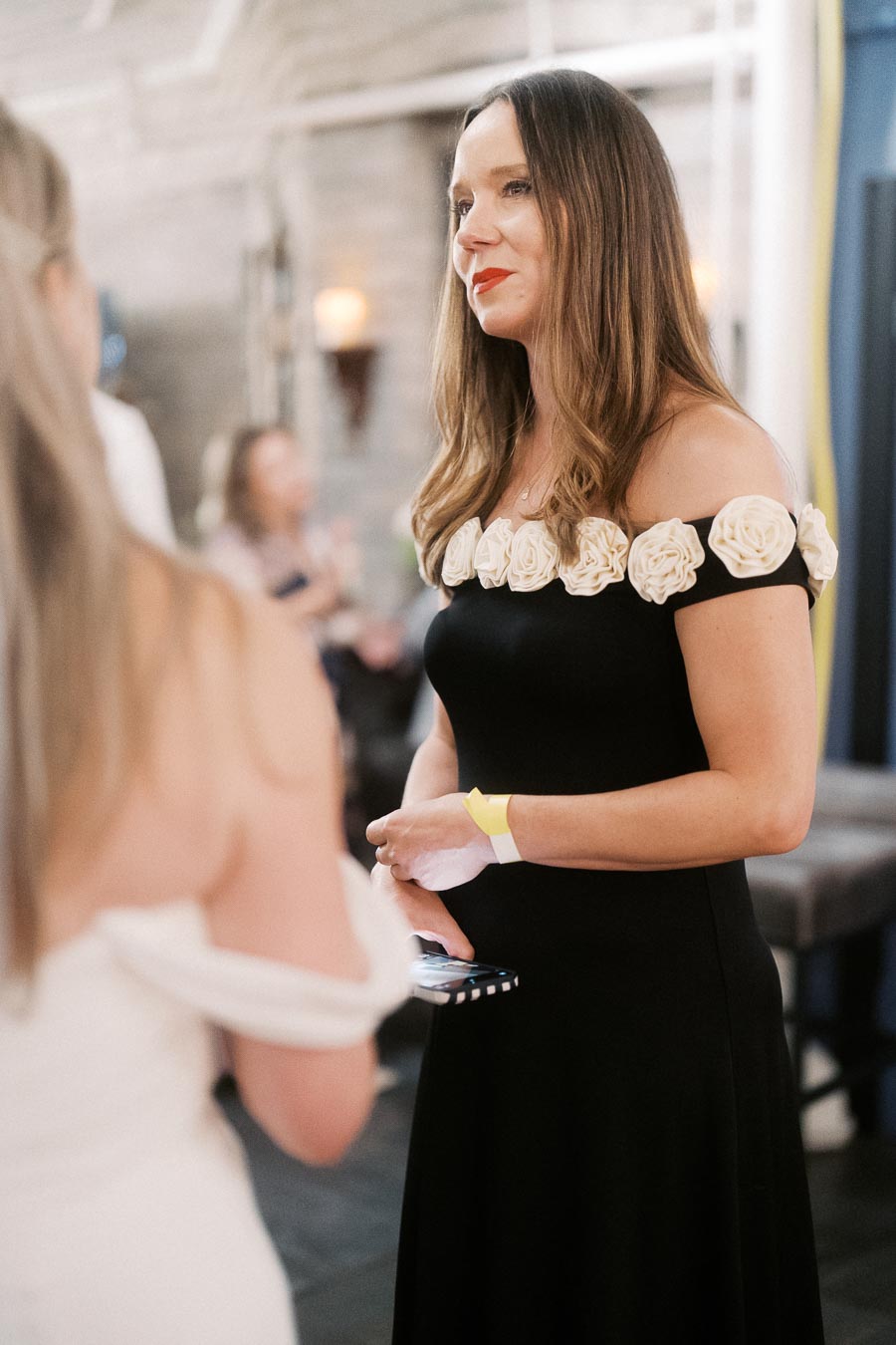 Woman in an elegant black dress with floral accents at a social event, engaged in conversation.