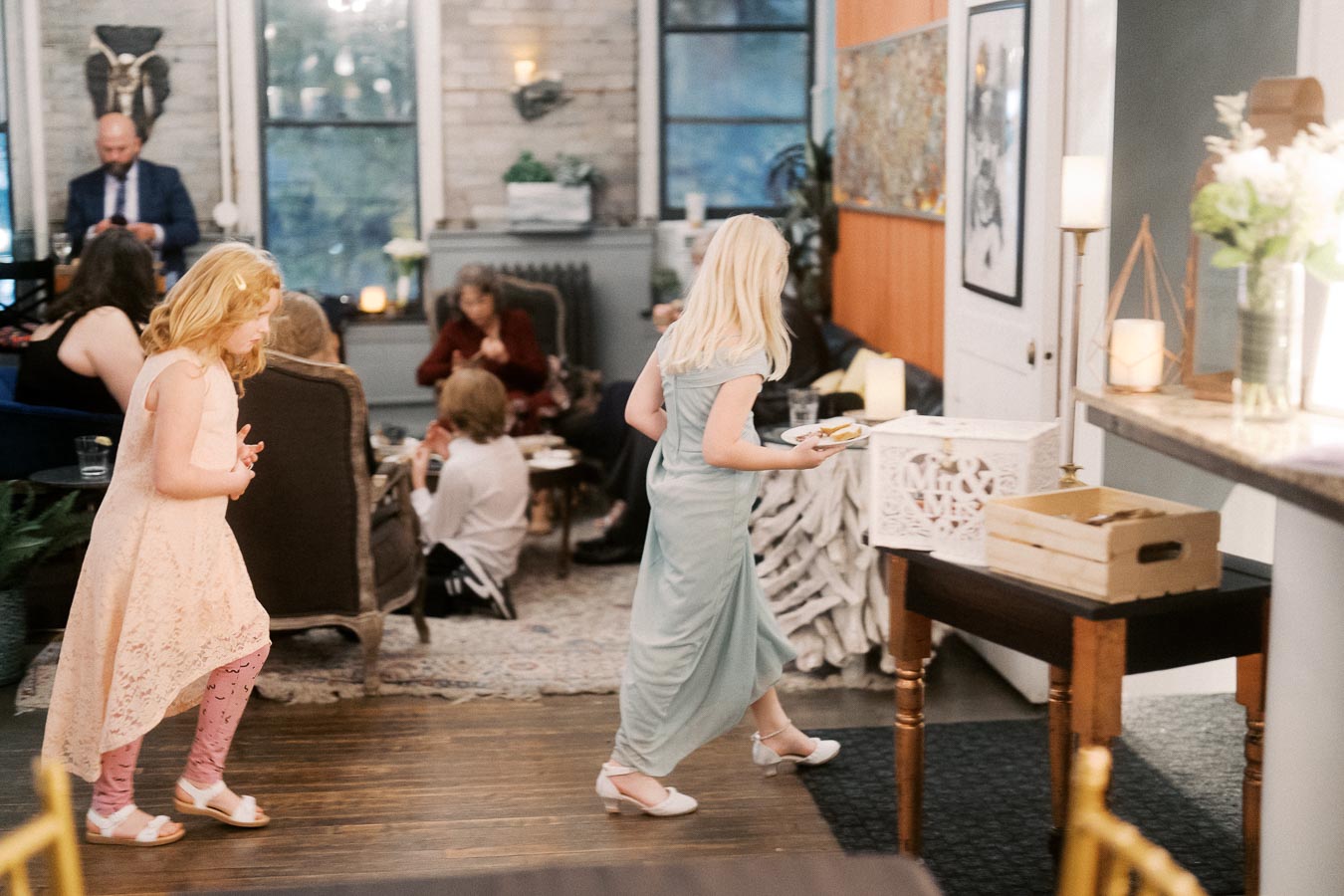 A cozy indoor gathering with young girls in dresses walking around, while adults sit and chat in the background. The warmly lit room features comfortable seating, elegant decor, and ambient lighting.