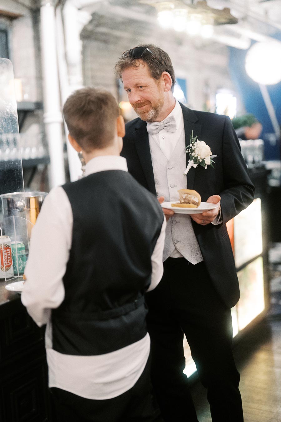 A man in a formal suit with a boutonniere conversing with a young boy at a wedding reception, holding a plate with a sandwich.
