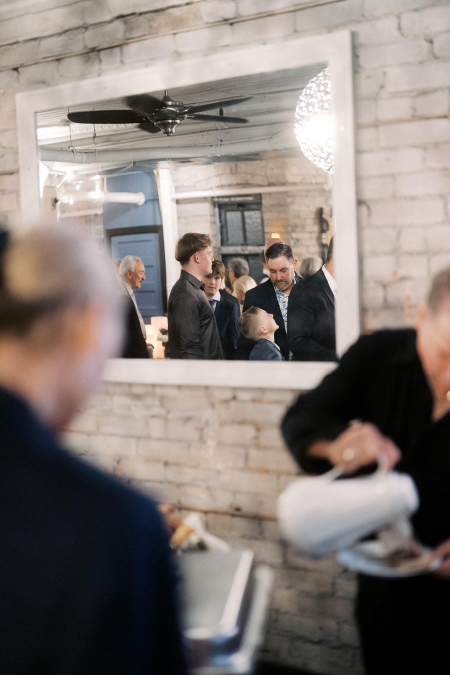Alt text: Group gathering reflected in a large mirror on a brick wall, with people engaged in conversation at an indoor event. Blurred foreground shows someone serving food from a buffet table.
