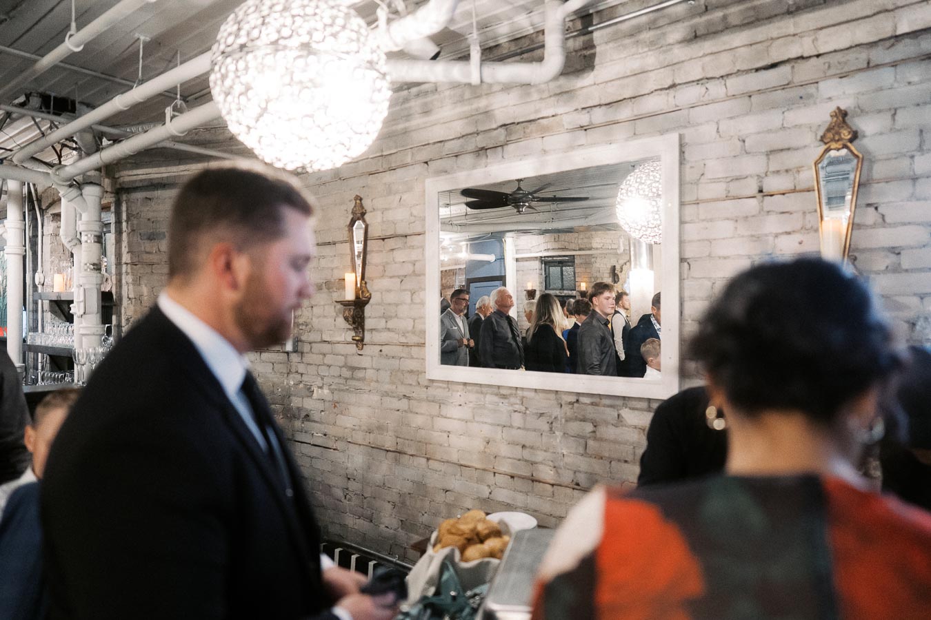 A stylish dinner event with guests socializing in a chic, industrial-style room featuring exposed brick walls, elegant mirrors, and a modern chandelier.