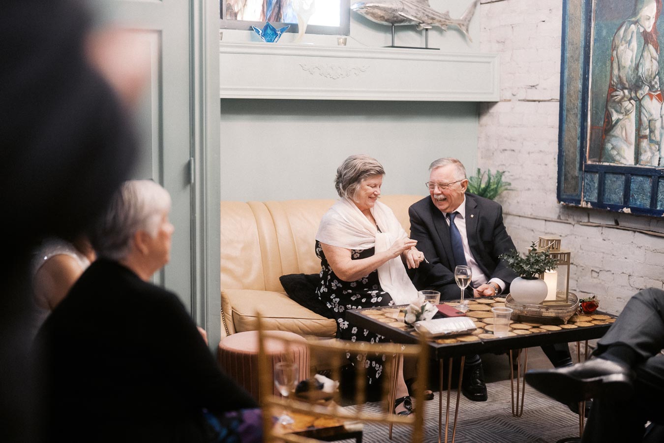 Elderly couple laughing and enjoying a cozy gathering in a stylish living room with a beige couch, decorative plants, and elegant artwork on the walls. A coffee table with drinks and decorative items sits in the foreground, creating a warm and inviting atmosphere.