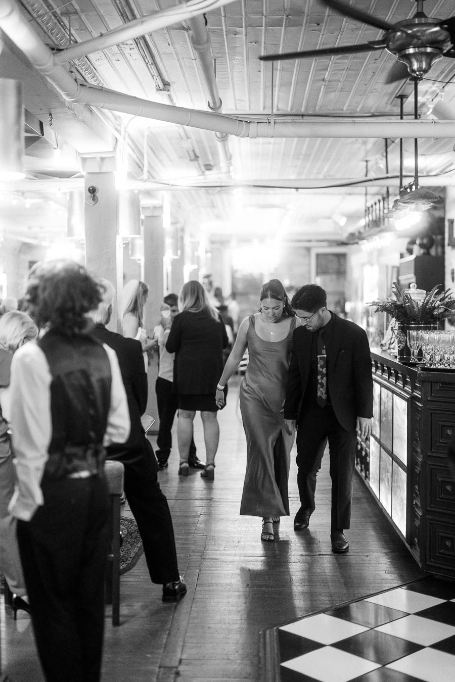 Black and white photo of a stylishly dressed couple walking hand in hand at an elegant indoor event venue, surrounded by other guests and soft ambient lighting.