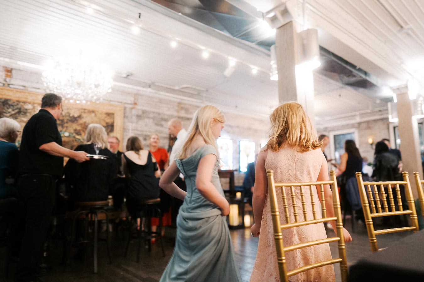 A lively indoor party scene with elegantly dressed guests socializing in a warmly-lit venue, featuring a chandelier, gold chairs, and a mix of casual and formal attire.