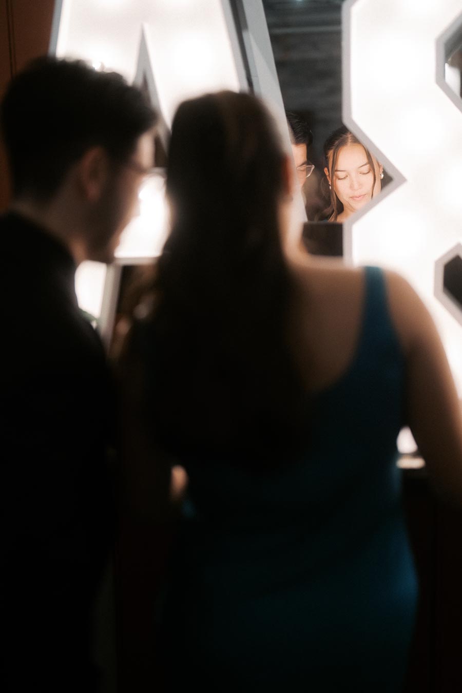Two people stand in front of an illuminated mirror, softly focused, creating a dreamy, intimate atmosphere as they share a moment of reflection together.