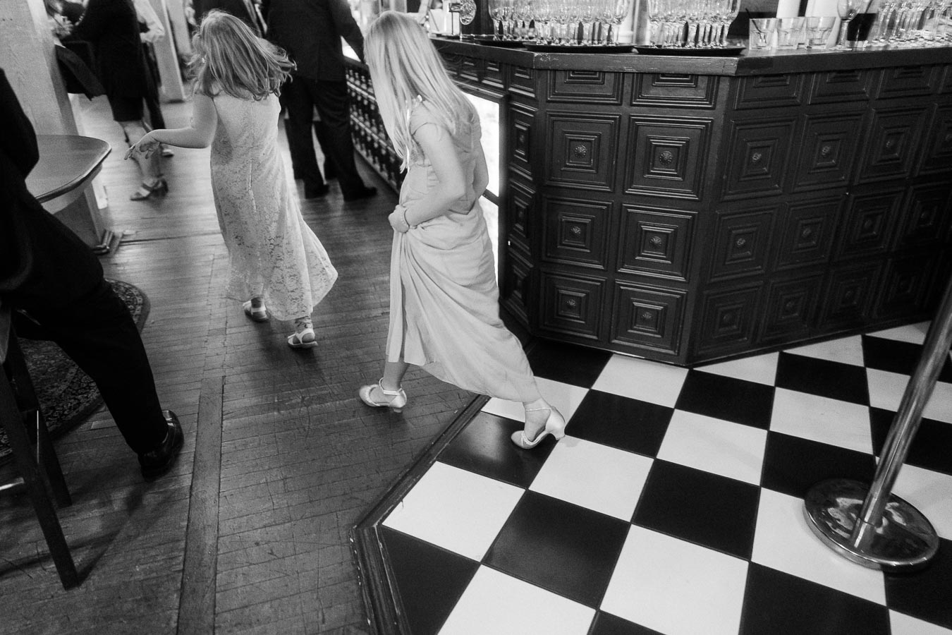 Black and white photo of two children in elegant dresses walking on a checkered floor at a formal event, with adults in suits visible in the background near a bar area.