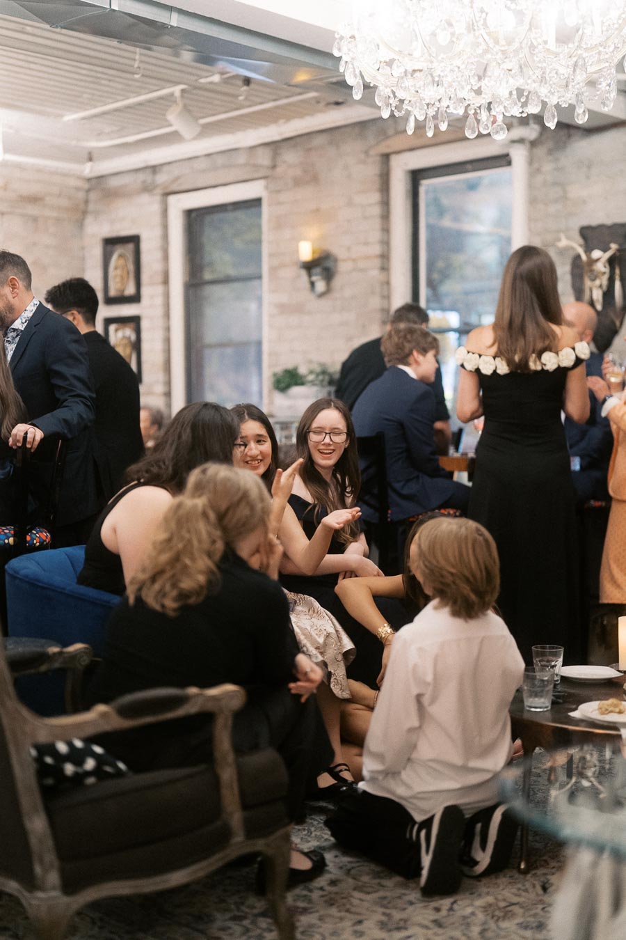 A lively gathering of people dressed elegantly, socializing in a stylish, well-lit room, featuring a chandelier, exposed brick walls, and artwork, creating a warm and inviting atmosphere.