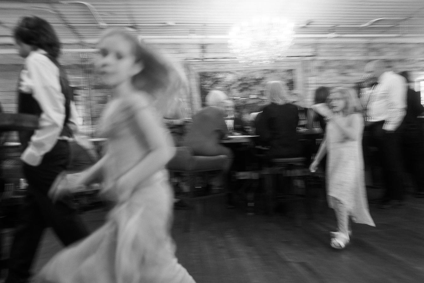 Blurred black and white image of children in motion at a social gathering, with seated adults in the background under a chandelier.