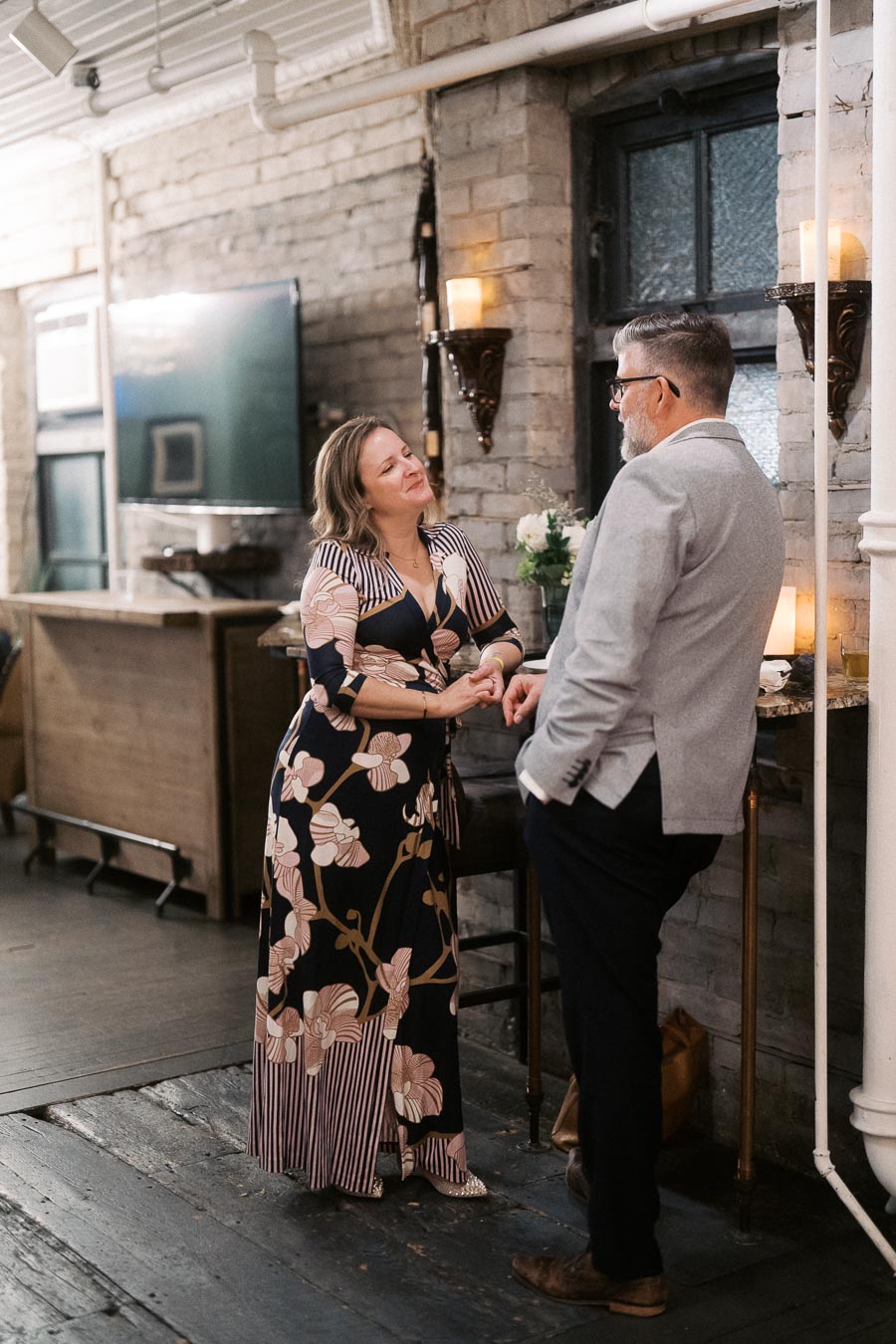 A woman in a floral dress and a man in a gray suit engage in a lively conversation in a cozy, dimly-lit room with exposed brick walls and candles.