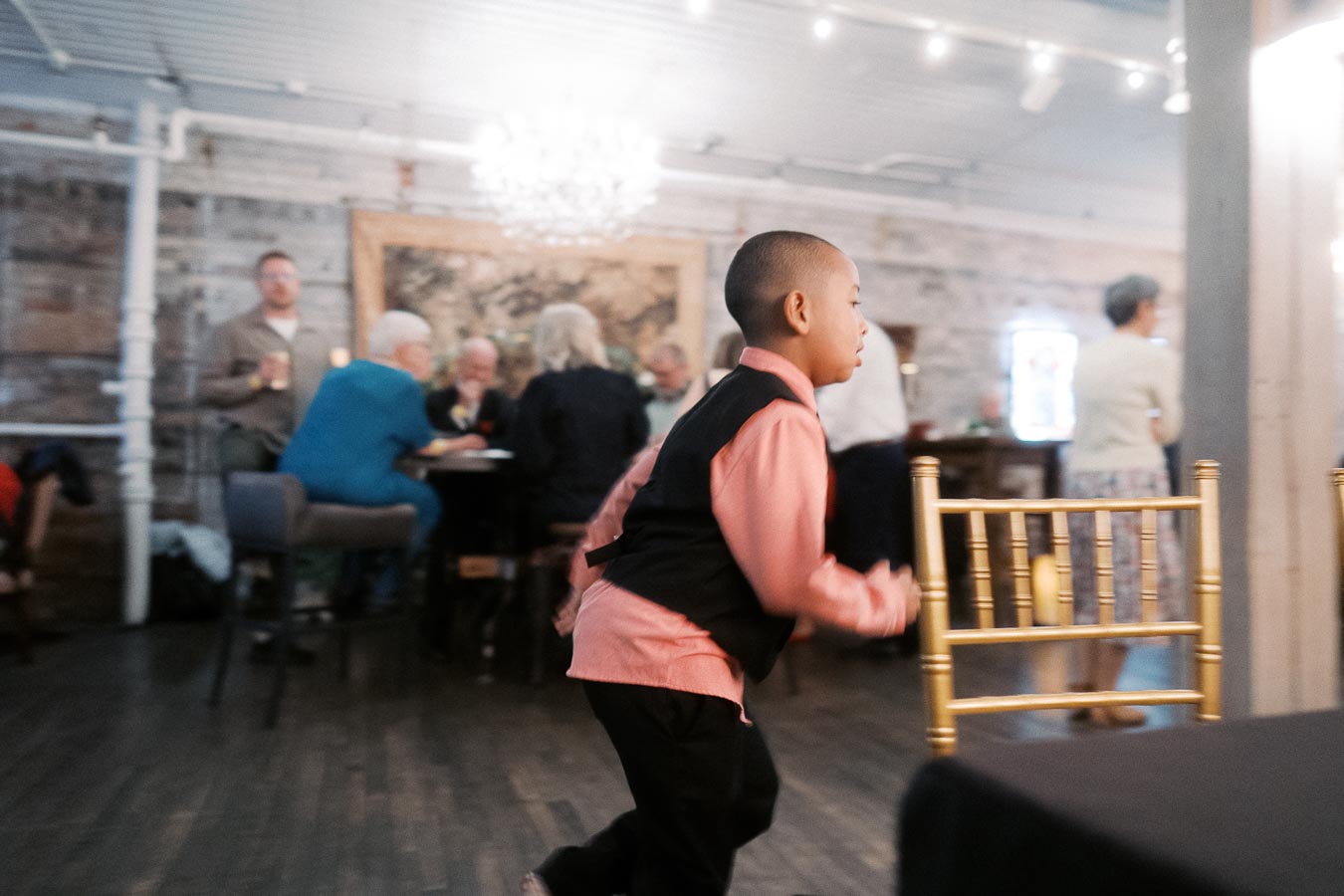 Young boy in a formal outfit running indoors at a lively social event, with adults chatting in the background and festive lighting creating a warm atmosphere.