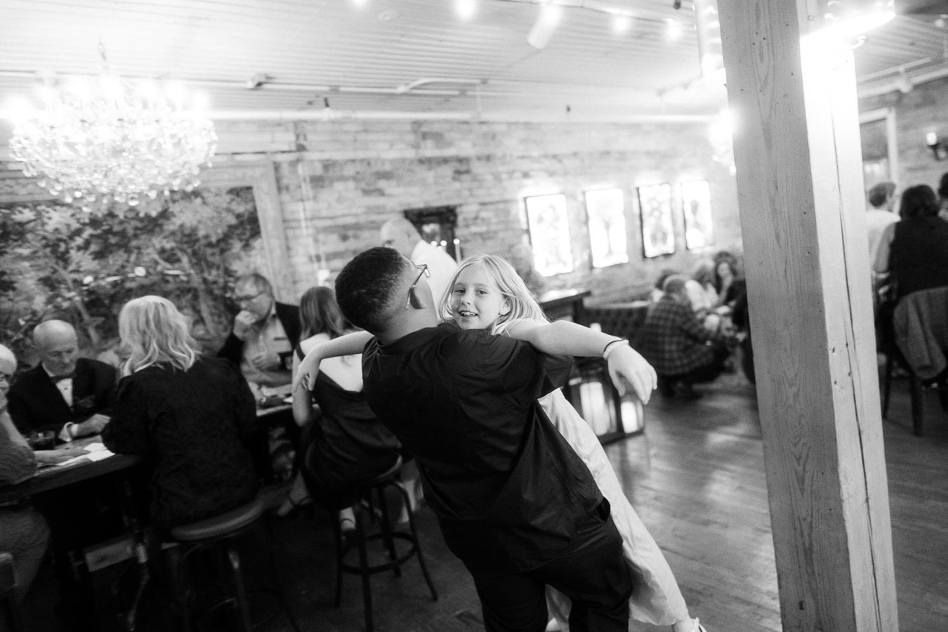 Black and white photo of a lively social gathering in a cozy venue with string lights and a chandelier. A young girl with blonde hair joyfully dances with a man in the foreground, while groups of people engage in conversation at tables in the background.