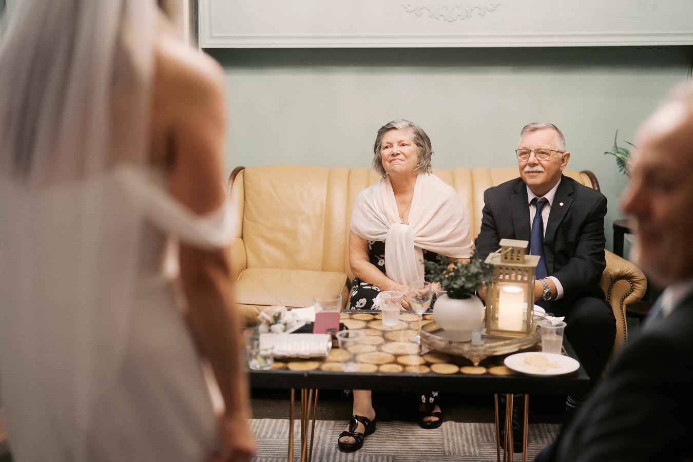 Elderly couple sitting on a sofa, attentively listening to a bride in a white gown at a wedding reception, with a decorative candle and plant on a table in a cozy room.