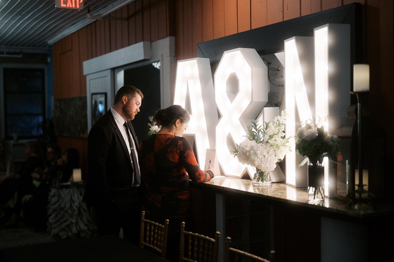 Couple standing by illuminated A&N letters at an elegant event, featuring floral arrangements and a warm, ambient setting.