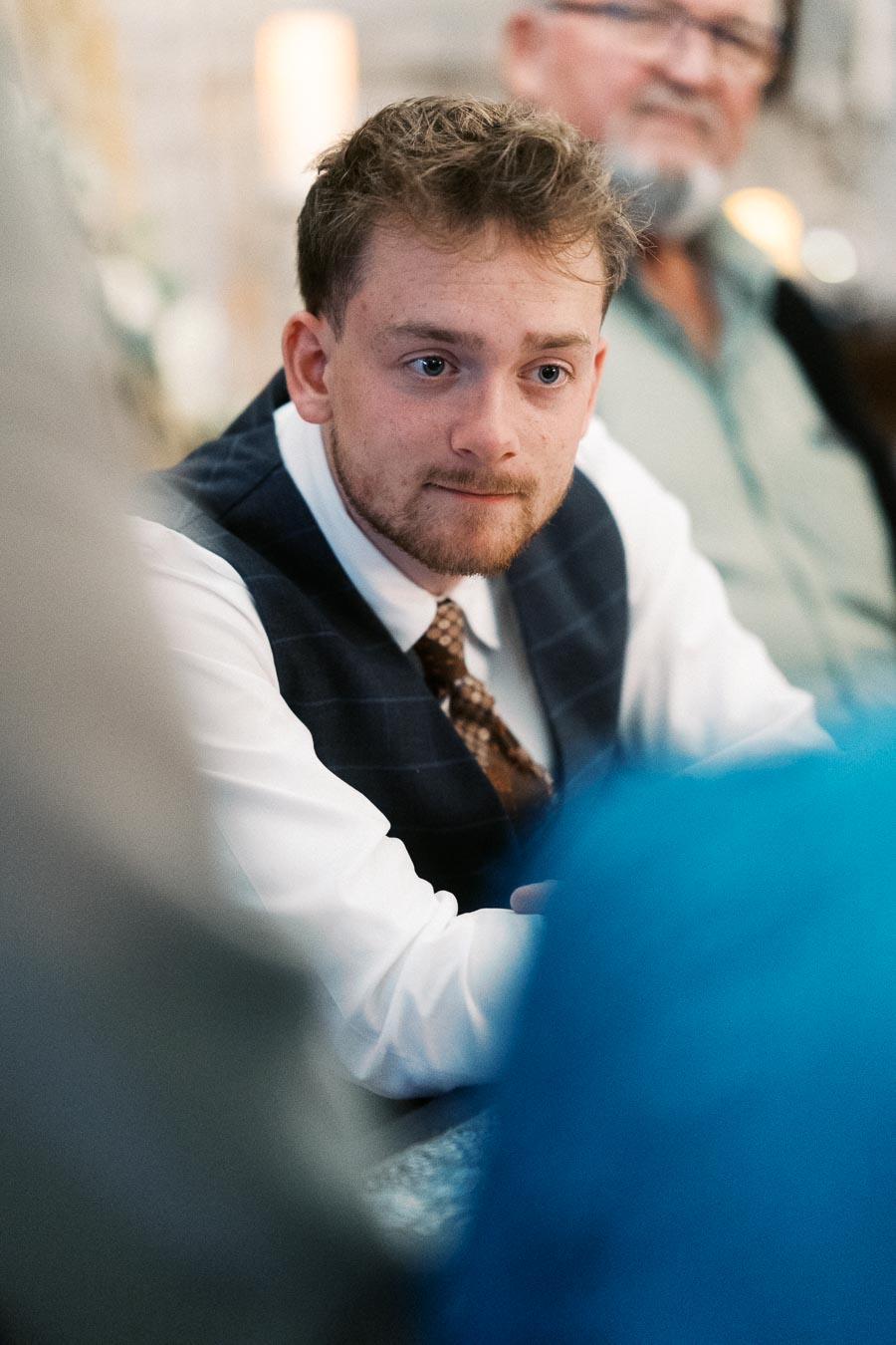 A young man in a formal outfit, including a vest and tie, is seated in a social setting, engaged in conversation. The background includes another person, slightly out of focus, in a casual shirt. The ambiance suggests a relaxed and intimate gathering.