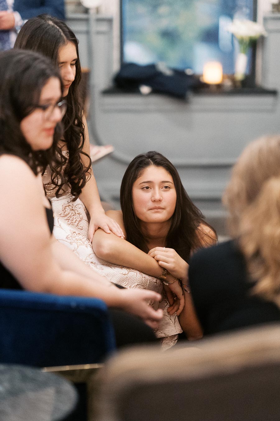 Group of young women in formal attire seated at an indoor event, one woman leaning on another's arm, with a blurred background featuring a lit candle and floral decoration.