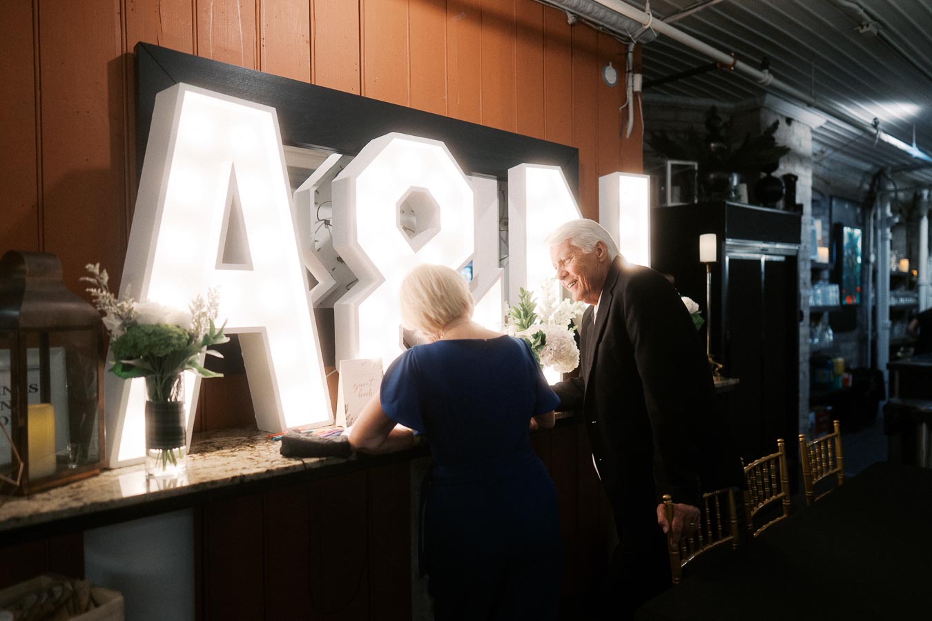 Elderly couple signing a guestbook at a wedding reception with illuminated A&N letters in the background.