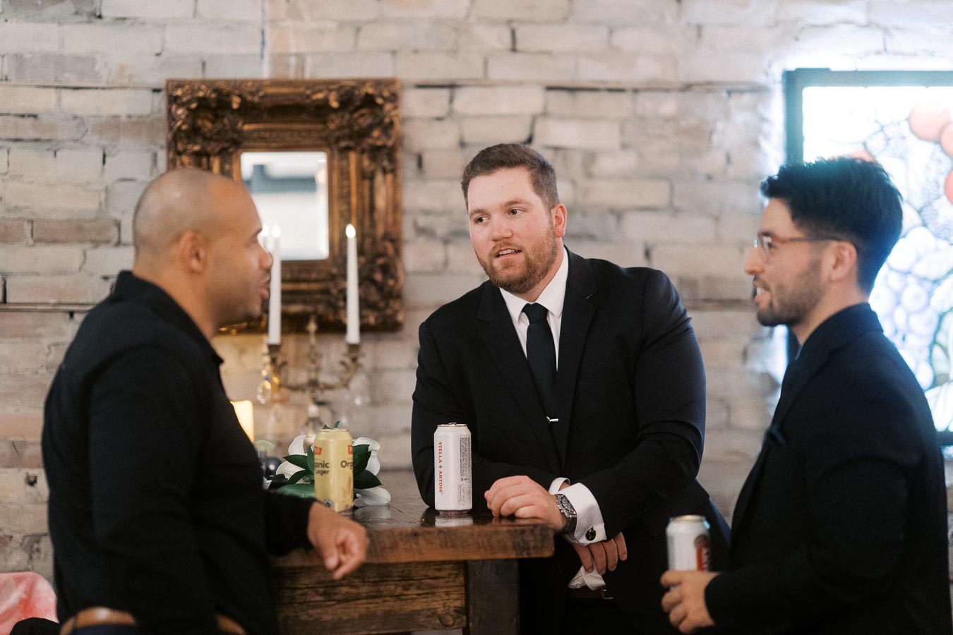Three men in formal attire engage in conversation at a rustic bar counter, holding cans of beverages.
