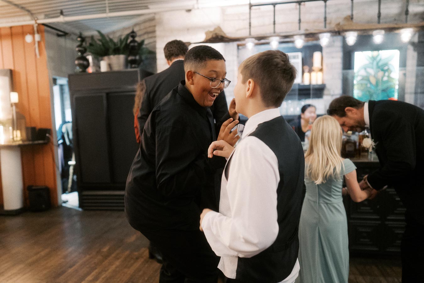 A group of well-dressed children and adults interact happily at a lively indoor event, with warm lighting and a modern decor background.
