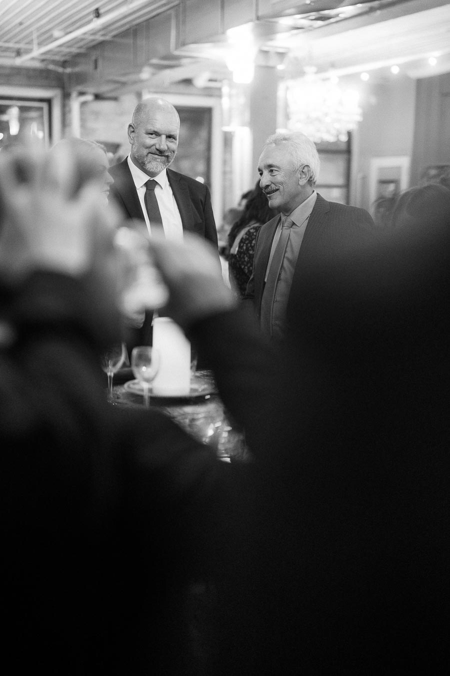 Black and white image of two men in business attire smiling and conversing at a formal event, surrounded by people, with soft lighting and elegant decor in the background.