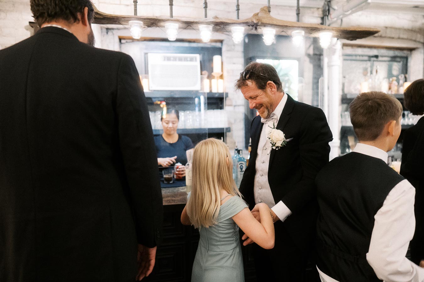 A man in a suit with a boutonnière smiling and holding hands with a young girl in a blue dress at a rustic bar setting, with bartenders in the background preparing drinks.