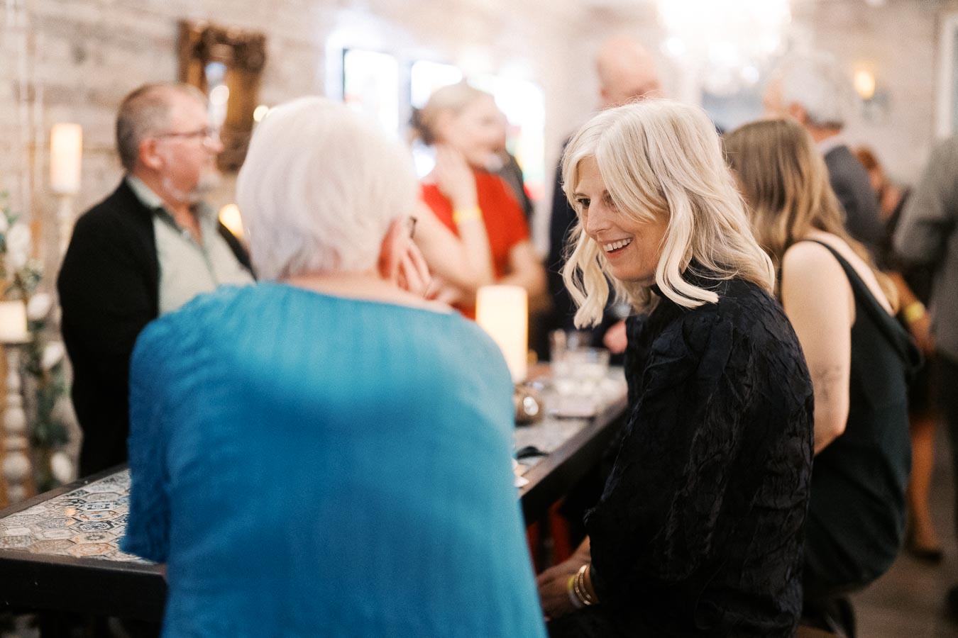 Group of people socializing at a lively indoor gathering, focusing on a woman with blonde hair smiling and interacting with friends around a table.
