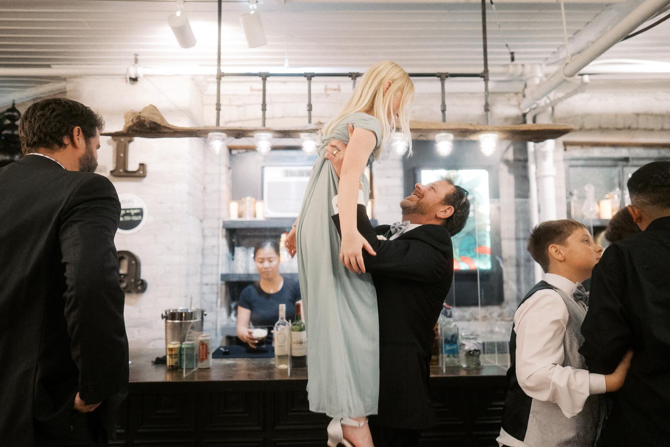A joyful moment at a lively gathering, with a man lifting a young girl in a pastel dress, surrounded by people enjoying a social event at a stylish, modern venue, featuring a bar and warm ambient lighting.