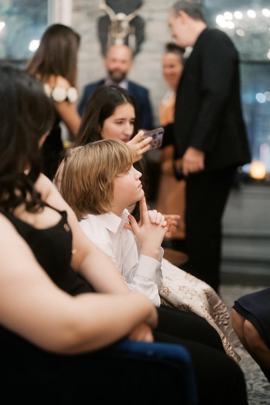 Group of people at a social gathering, including a child in a white shirt appearing thoughtful, with others engaged in conversation and capturing the moment with a smartphone in a warmly lit room.