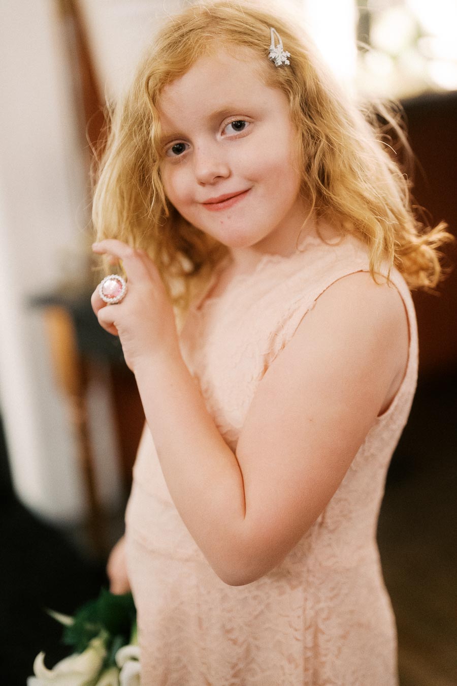 Young girl with curly blonde hair smiling and showing off a decorative ring, wearing a light pink lace dress and hair accessory, indoors.