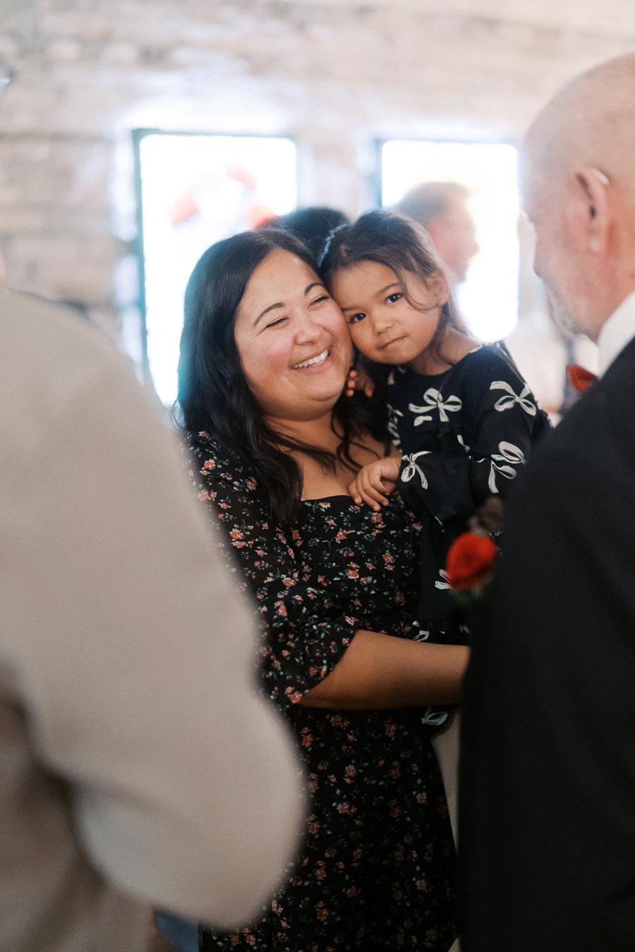 A woman holding a child and smiling warmly in a softly lit room, surrounded by other people during a social gathering.