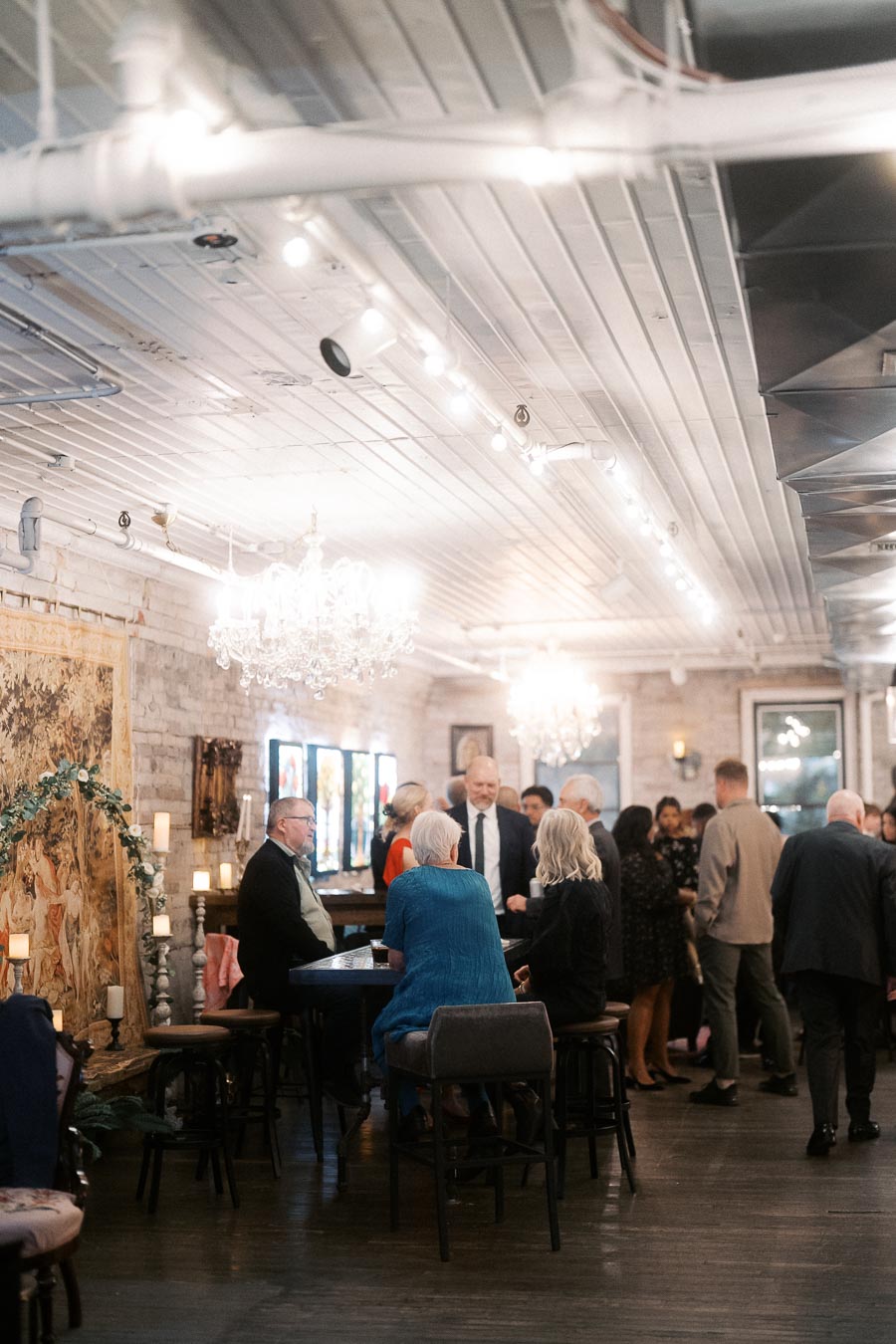Elegant social gathering in a stylishly decorated venue with people mingling around tables, under chandeliers in a warmly lit room featuring brick walls and artistic tapestries.
