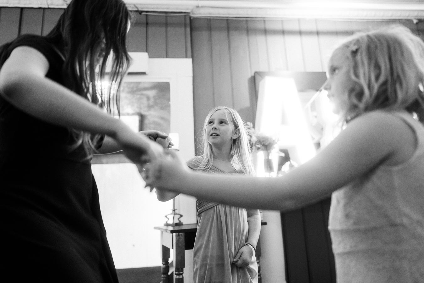 Black and white image of three young girls dancing together indoors, surrounded by a cozy setting with bright lights in the background.