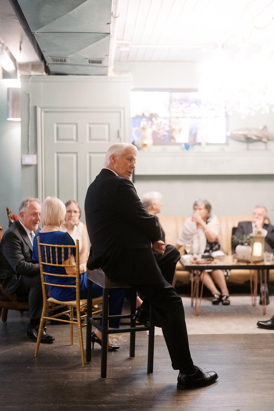 Elegantly dressed elderly gentleman sitting at a casual indoor gathering, conversing with a group of people in a warmly lit room.