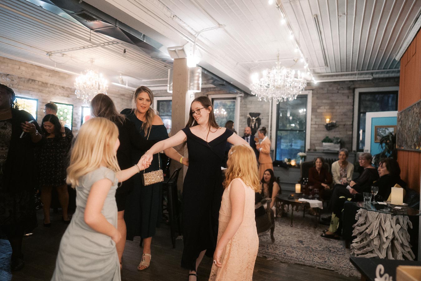 A lively indoor party scene featuring people of various ages interacting and dancing. Chandeliers hang from the ceiling, and guests are seated and standing in a warmly lit, stylishly decorated room.
