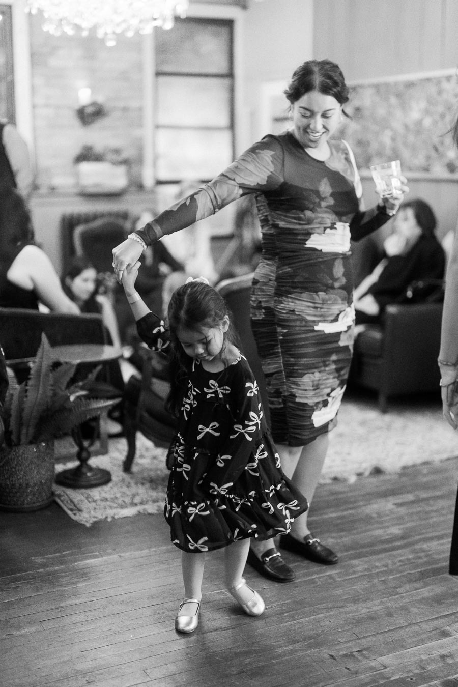 Woman and young girl dancing together at a social gathering, both smiling and holding hands. The room has a cozy atmosphere with several people seated in the background, and a decorative plant visible in the foreground.